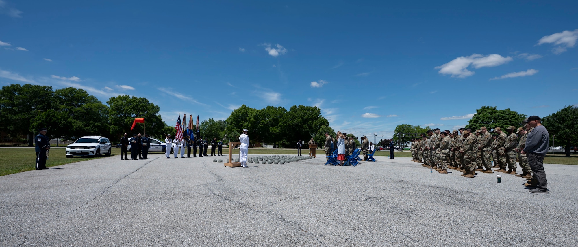A wide shot depicts an audience, right, observing a military ceremony, left, under a bright blue sky.
