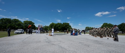 A wide shot depicts an audience, right, observing a military ceremony, left, under a bright blue sky.