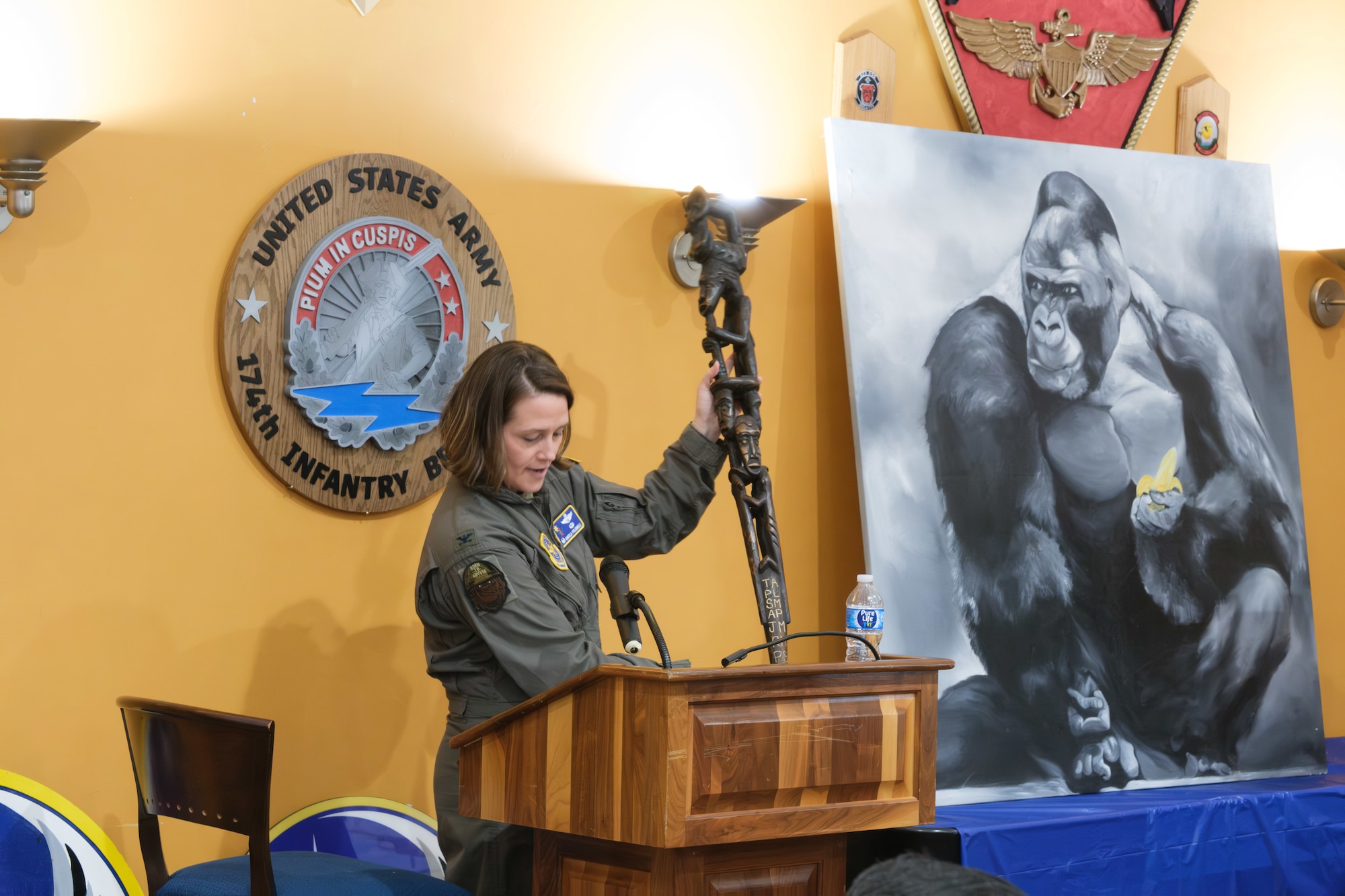 A woman in a flight suit stands at a podium and holds a ceremonial mace. A U.S. Army 174th Infantry Brigade seal and a painting of a gorilla are in the background.