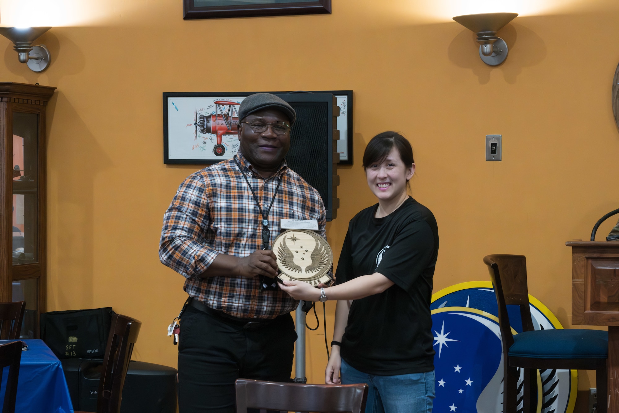 A woman presents a plaque to a man in front of an orange wall during an awards presentation.