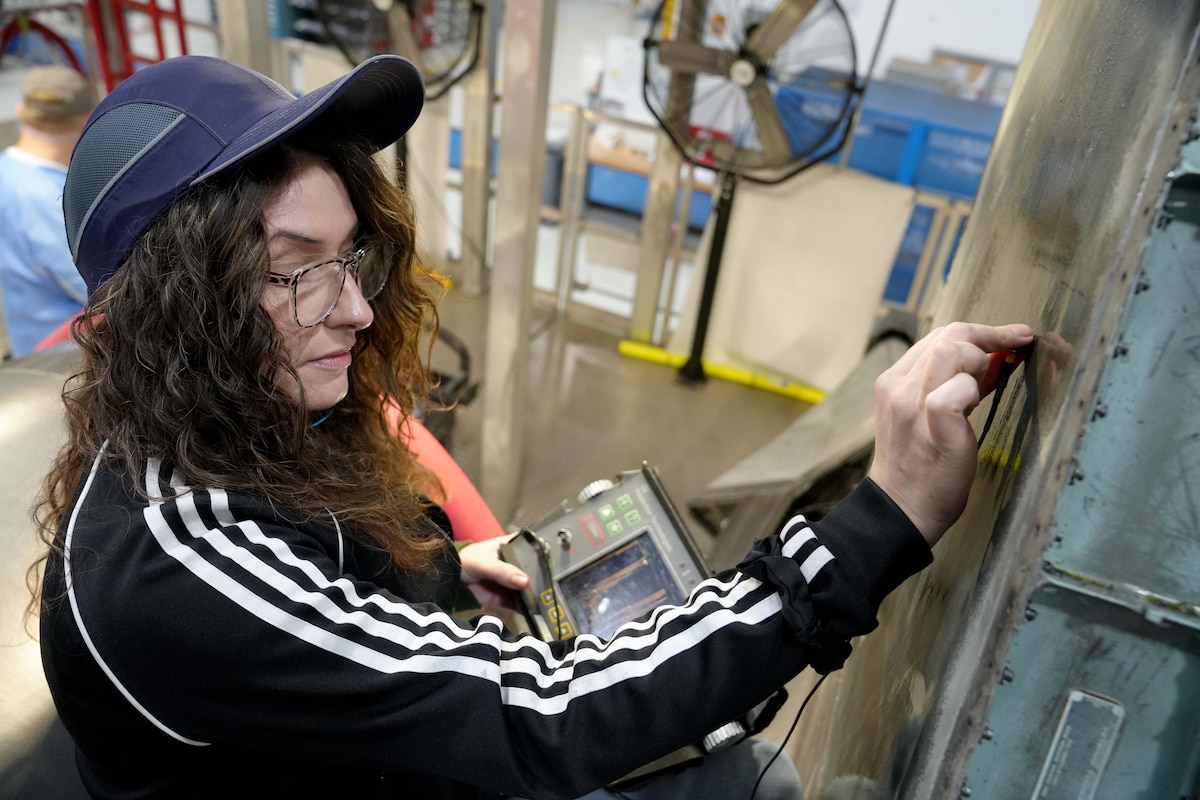 Photo shows woman using a machine to perform measurements on a large piece of metal.