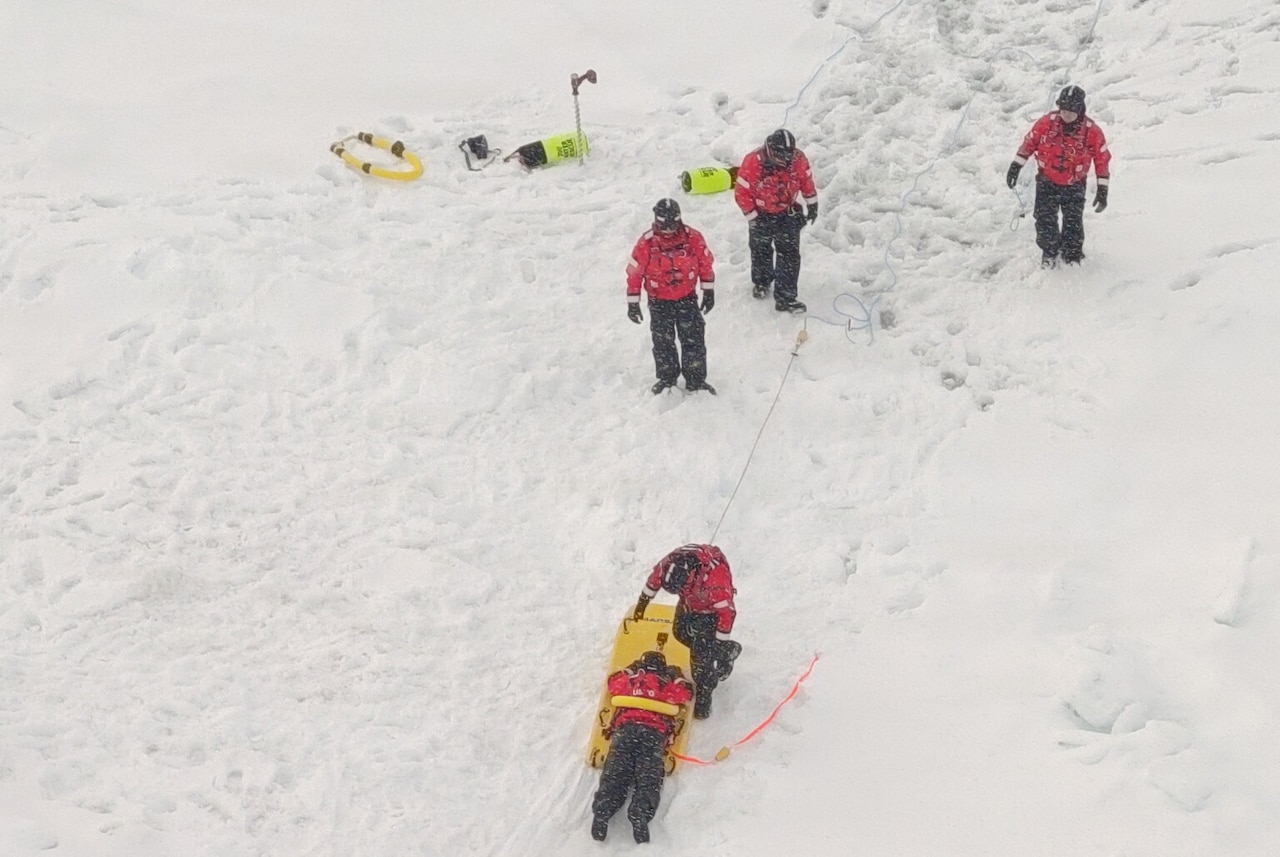 Three people in winter attire walk through the snow toward another person in similar attire who is putting a simulated patient on a stretcher.