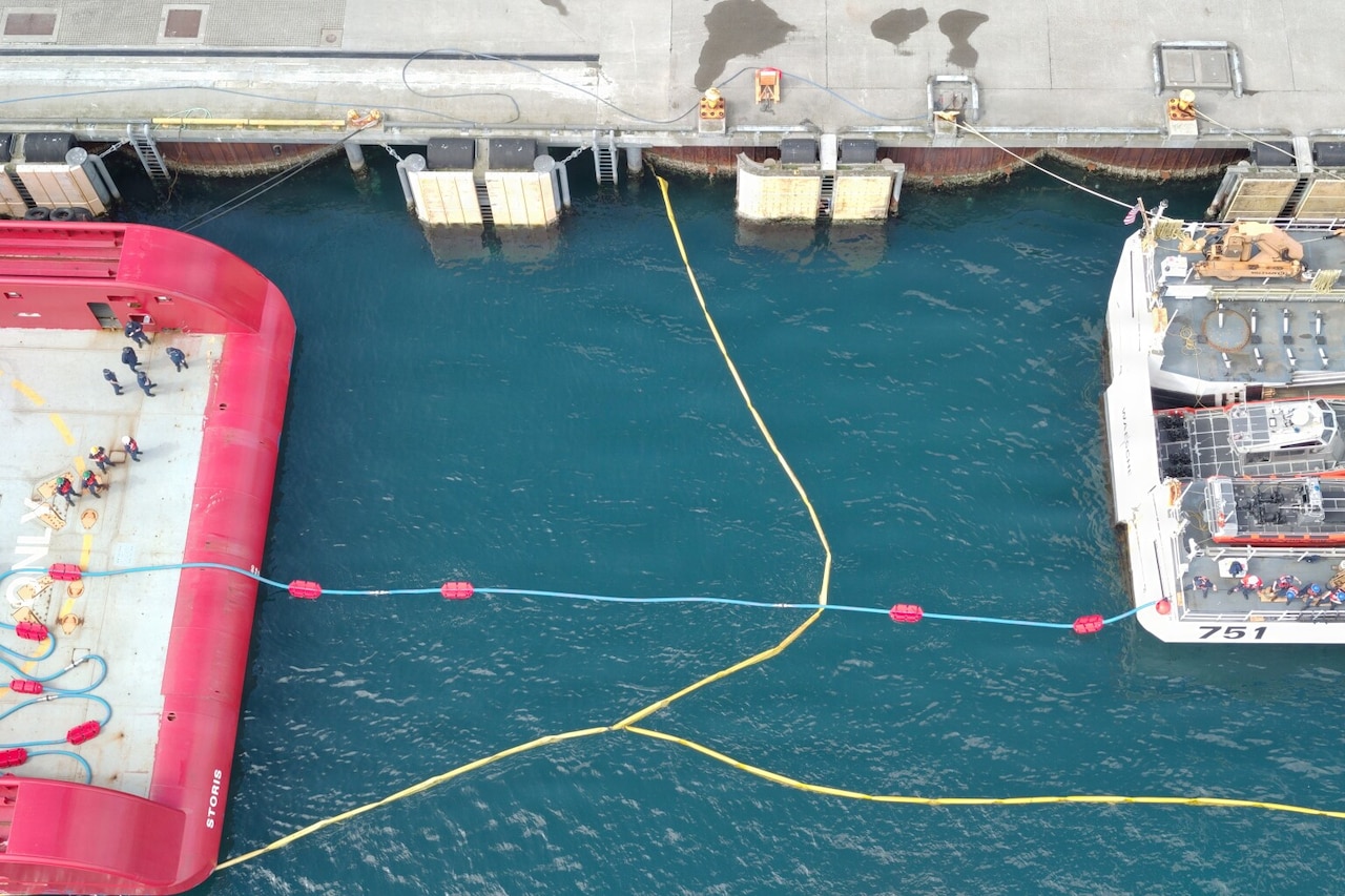 An aerial view of dozens of people standing at the stern of two ships as they connect fuel lines for a transfer; both ships are moored at a port.