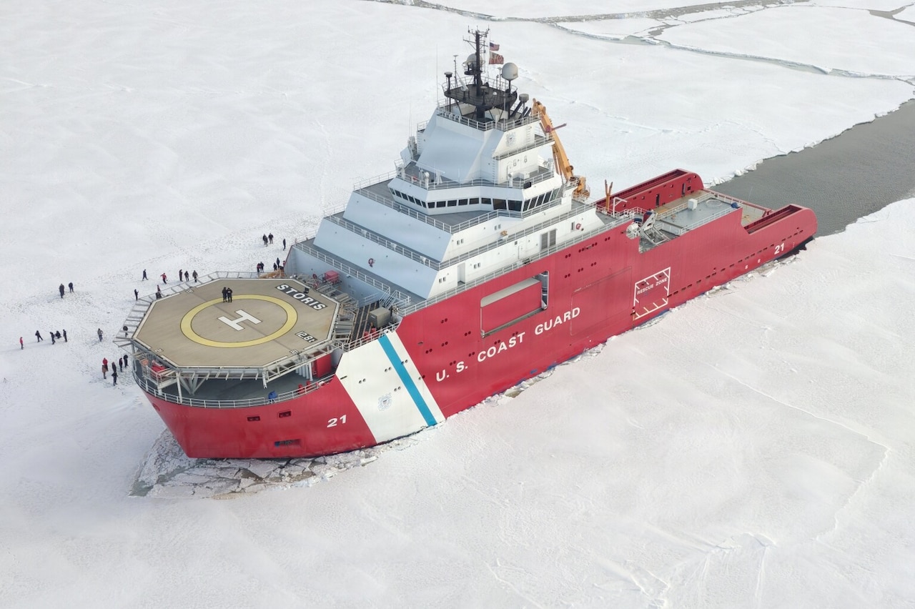 A large ship sits in the icy water with an open patch of water behind it; there are several people standing on the ice beside the ship.