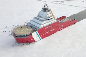 A large ship sits in the icy water with an open patch of water behind it; there are several people standing on the ice beside the ship.