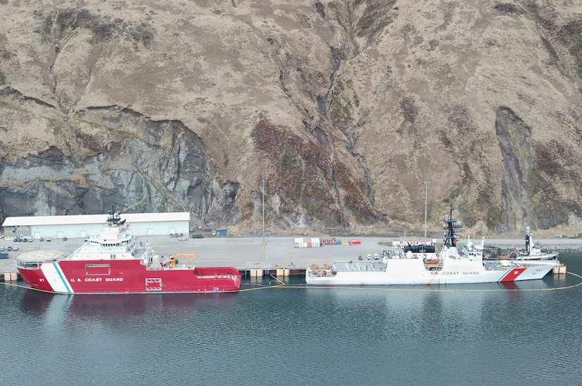 Two ships are moored at a port with both sterns facing each other for a fuel transfer; there are mountains in the background.