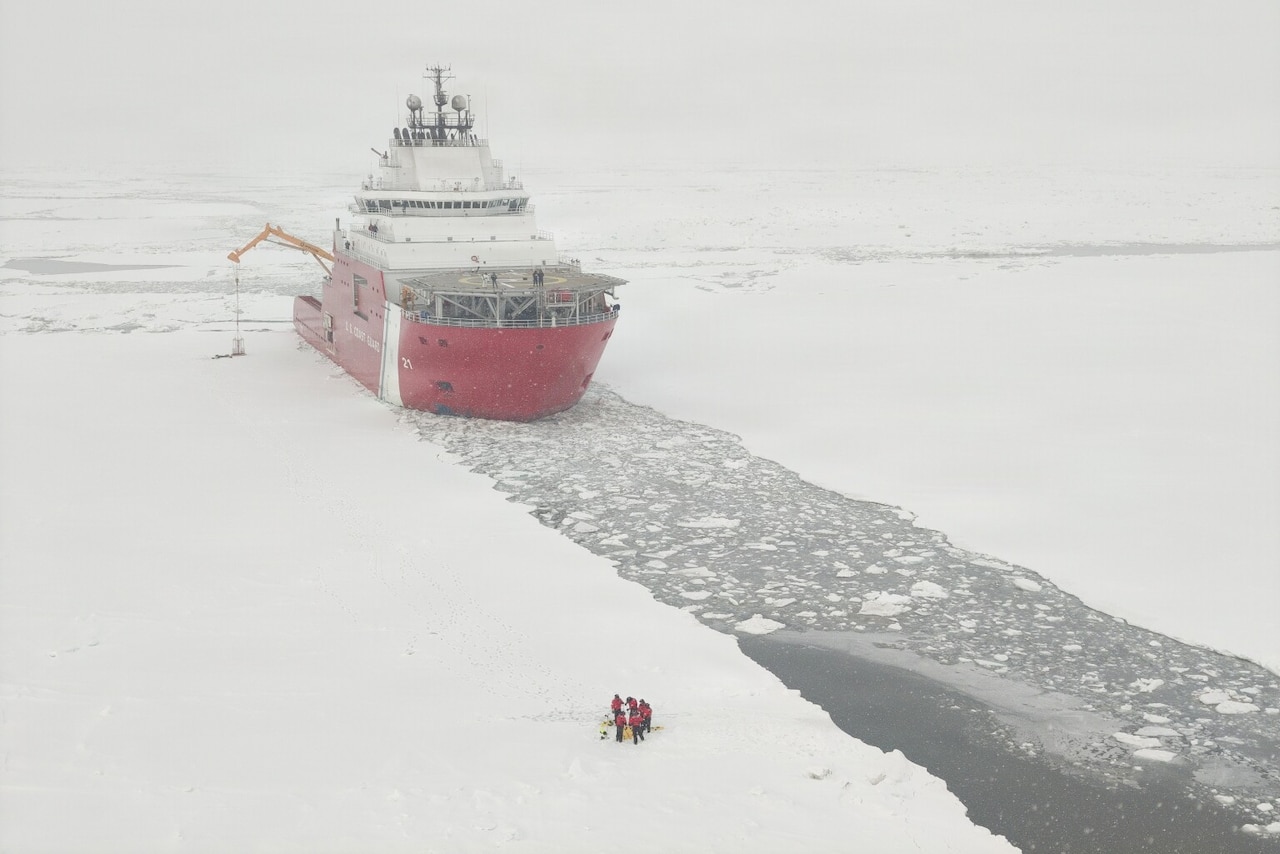 A large ship sits in icy water as a group of people in winter attire conduct training on the ice.