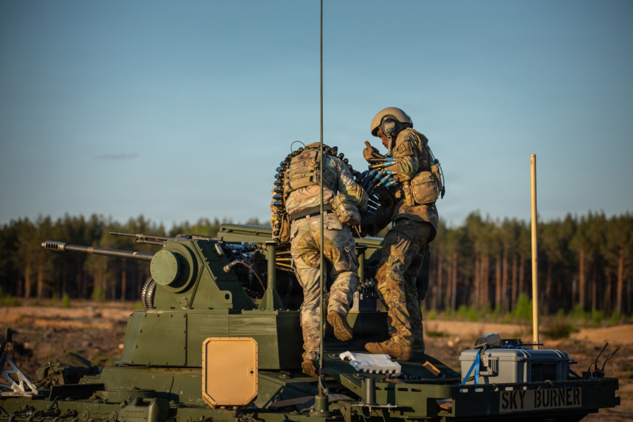 Two people wearing camouflage military uniforms load ammunition onto a military cannon.