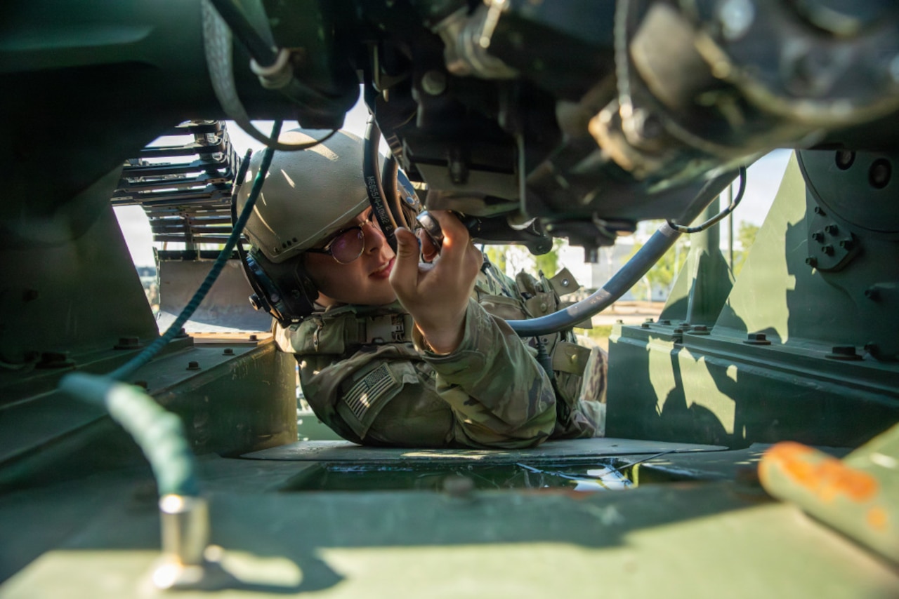 A person wearing a camouflage military uniform kneels down to work on a military cannon.