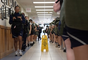 U.S. Airmen walk in a formation inside a hallway while carrying duffle bags.