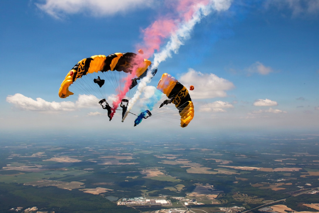 Three soldiers pose with golden and black parachutes in a blue sky while spraying red, white and blue smoke.