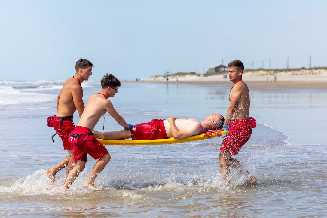 Three Marine Corps lifeguards carry another lifeguard on a stretcher out of the ocean under a blue sky.