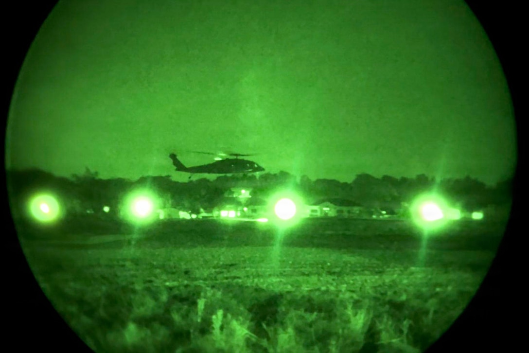 A helicopter flies over a wooded area as four bright lights shine on the ground as seen through a night vision lens.