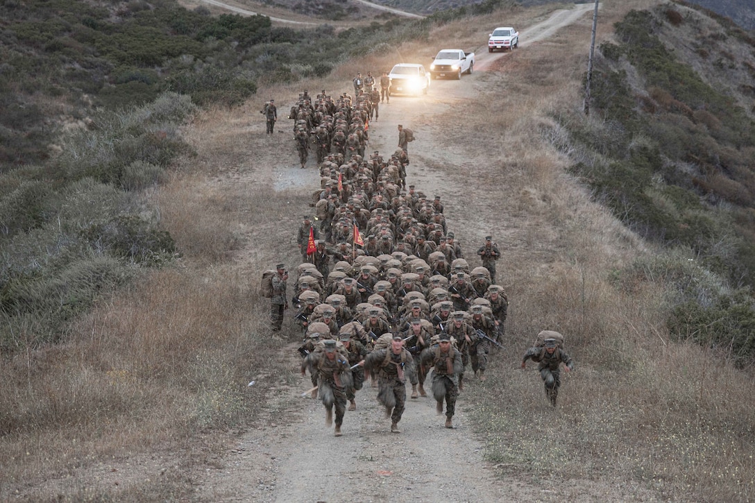 Dozens of Marines in tactical gear climb a dirt hill with three white pickup trucks in the background.