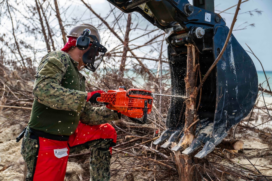 A service member surrounded by branches and debris on a beach uses a chainsaw to cut a tree branch stuck in a large backhoe bucket.