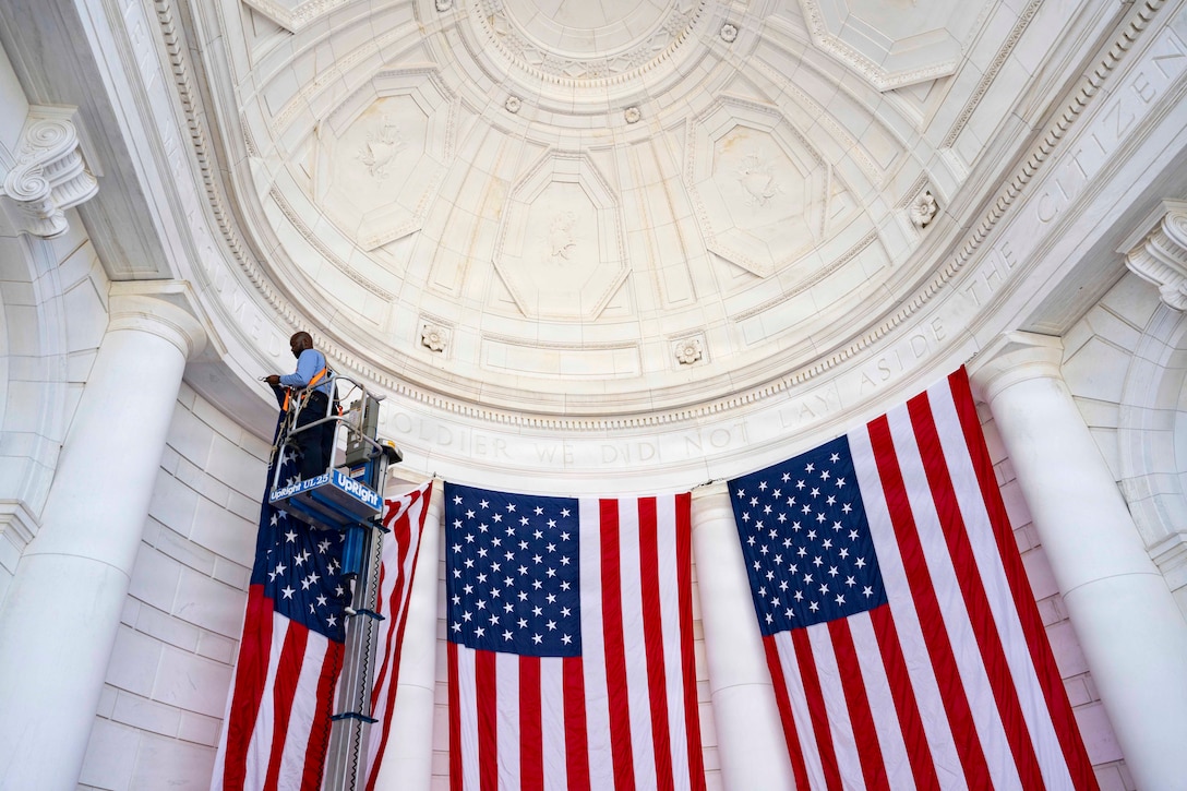 A person in a boom lift hangs an American flag high on the wall just under the ceiling of an ampitheather next to two other American flags.