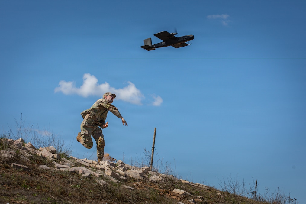 A soldier throws a small drone from the top of a hill, with a blue sky and sprinkle of white clouds in the background.