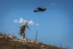 A soldier throws a small drone from the top of a hill, with a blue sky and sprinkle of white clouds in the background.