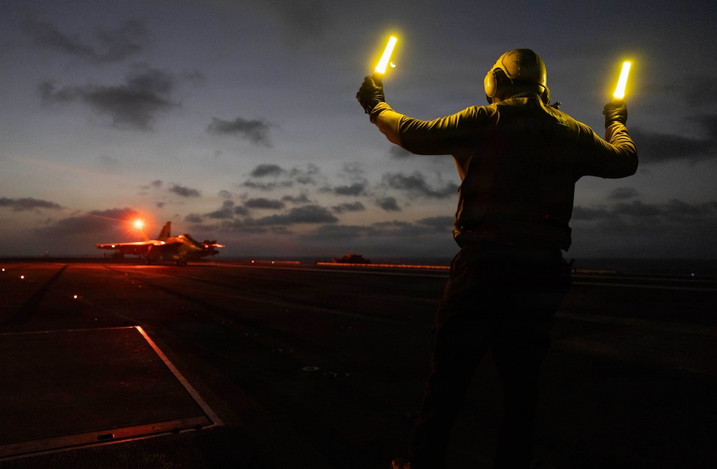 A person holds up a light stick in each hand, with their back to the camera, and faces a jet more than a hundred yards away on the deck of an aircraft carrier as the sun sets.
