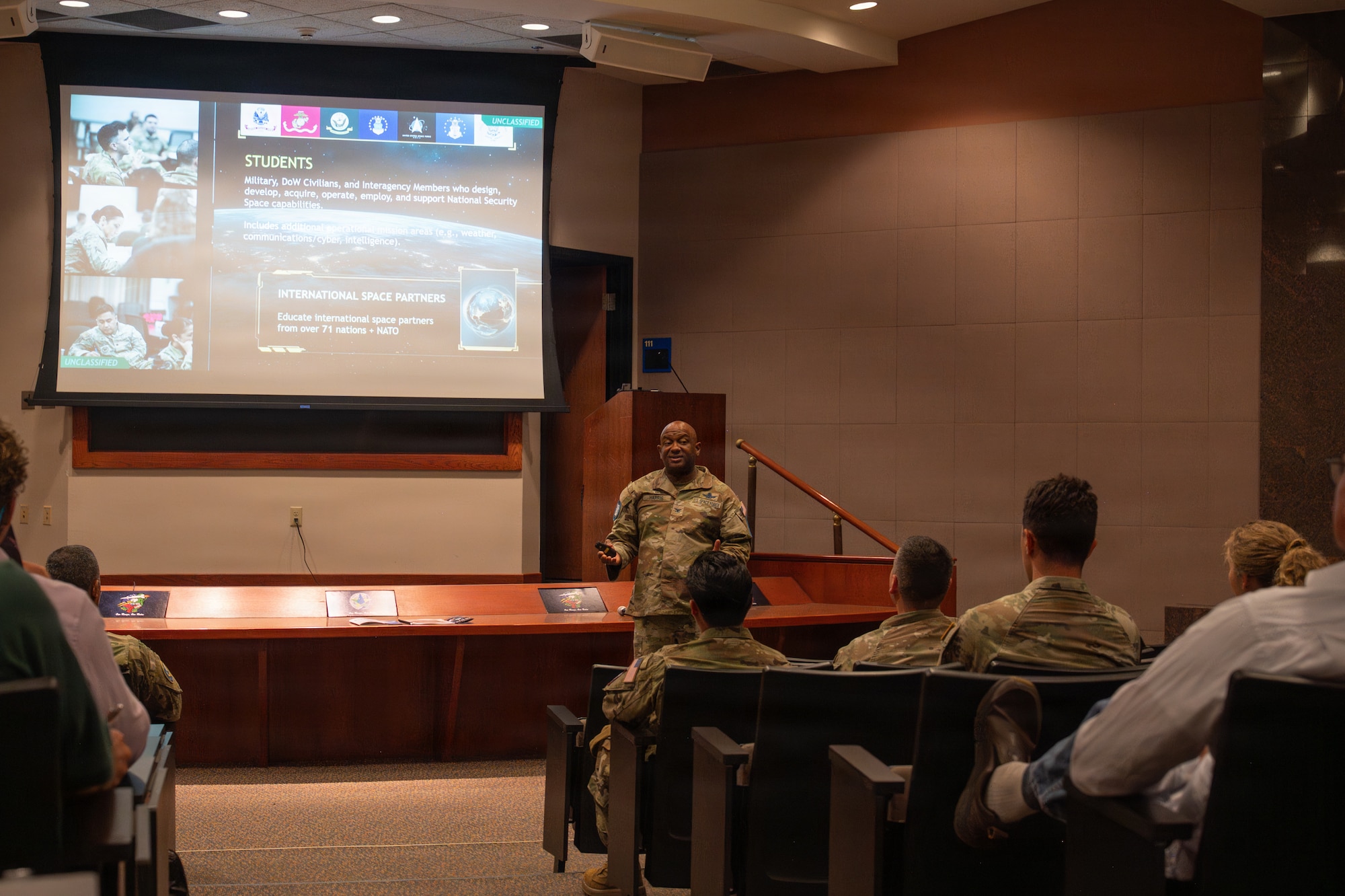 A person stands and speaks to a group of people sitting down inside an auditorium.