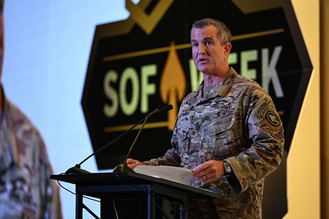 A man wearing a military camouflage uniform stands indoors behind a lectern speaking into a microphone.