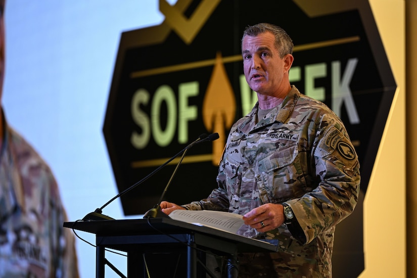 A man wearing a military camouflage uniform stands indoors behind a lectern speaking into a microphone.