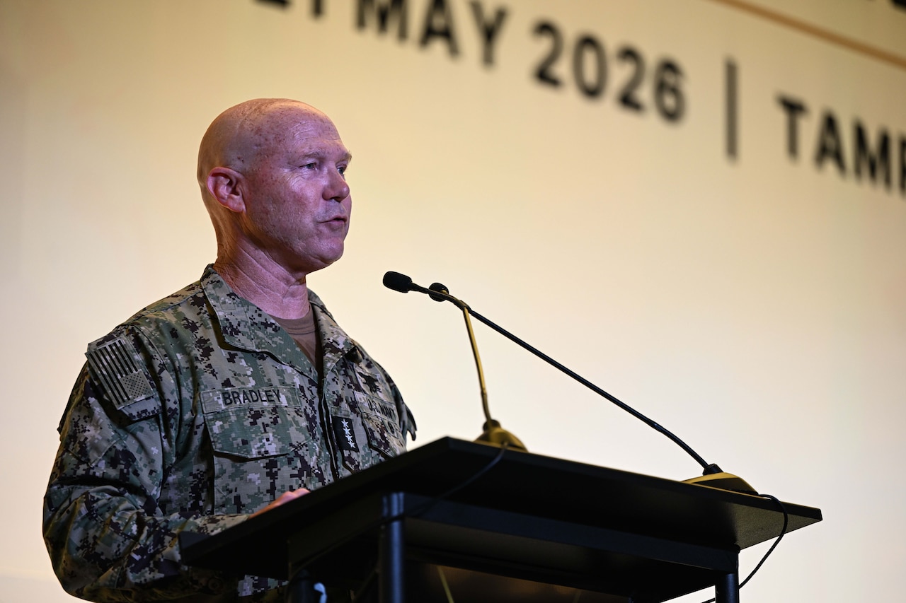 A man wearing a military camouflage uniform stands indoors behind a lectern speaking into a microphone.