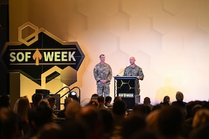 Two men wearing camouflage military uniforms stand indoors on a stage behind a lectern looking out at a large crowd. To the left of the men is a large sign that reads, "SOF Week."