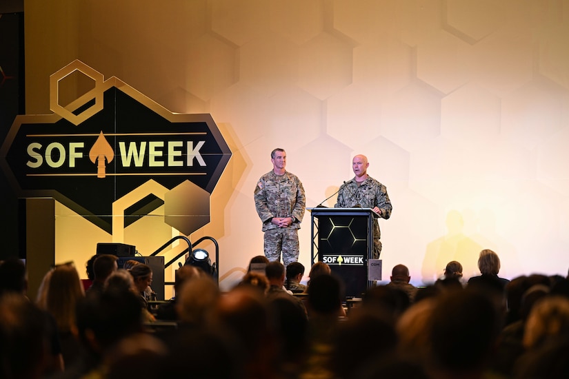 Two men wearing camouflage military uniforms stand indoors on a stage behind a lectern looking out at a large crowd. To the left of the men is a large sign that reads, "SOF Week."