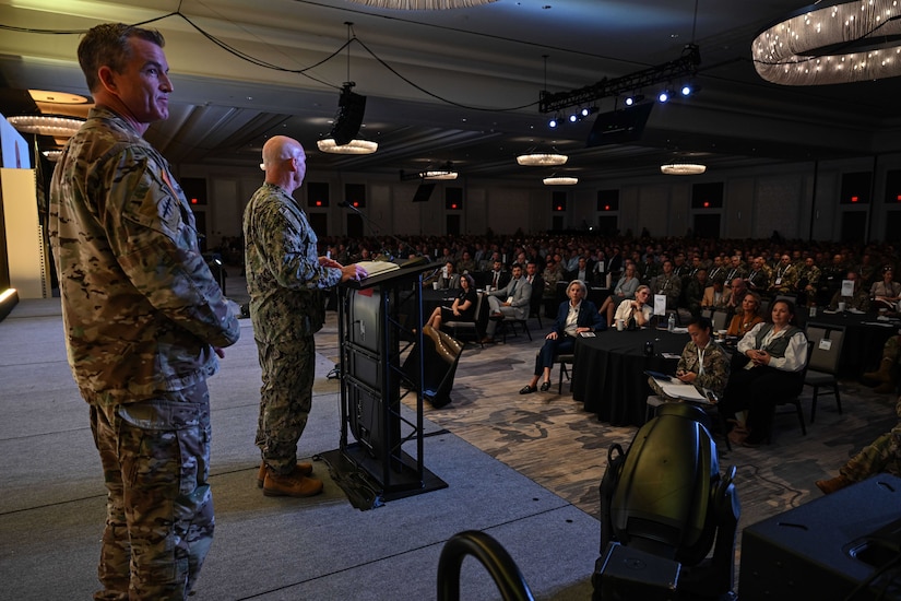 Two men in camouflage military uniforms stand indoors on a stage behind a lectern looking out at a large crowd.