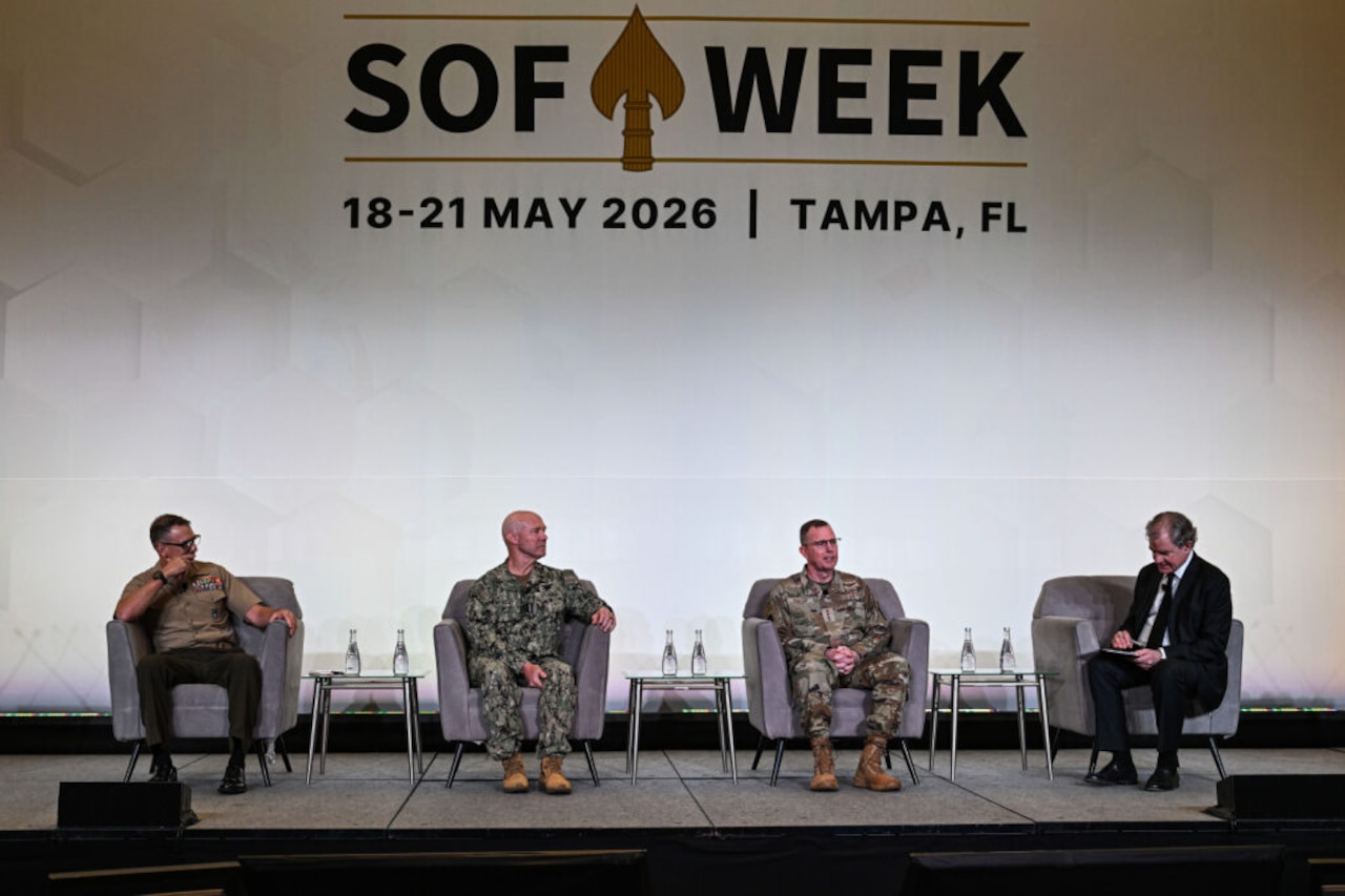 Four men, three of them in camouflage military uniforms and one wearing a business suit, sit in chairs indoors on a stage, having a discussion. Behind them on the wall is a sign that reads "SOF Week 18-21 May 2026, Tampa, Florida."