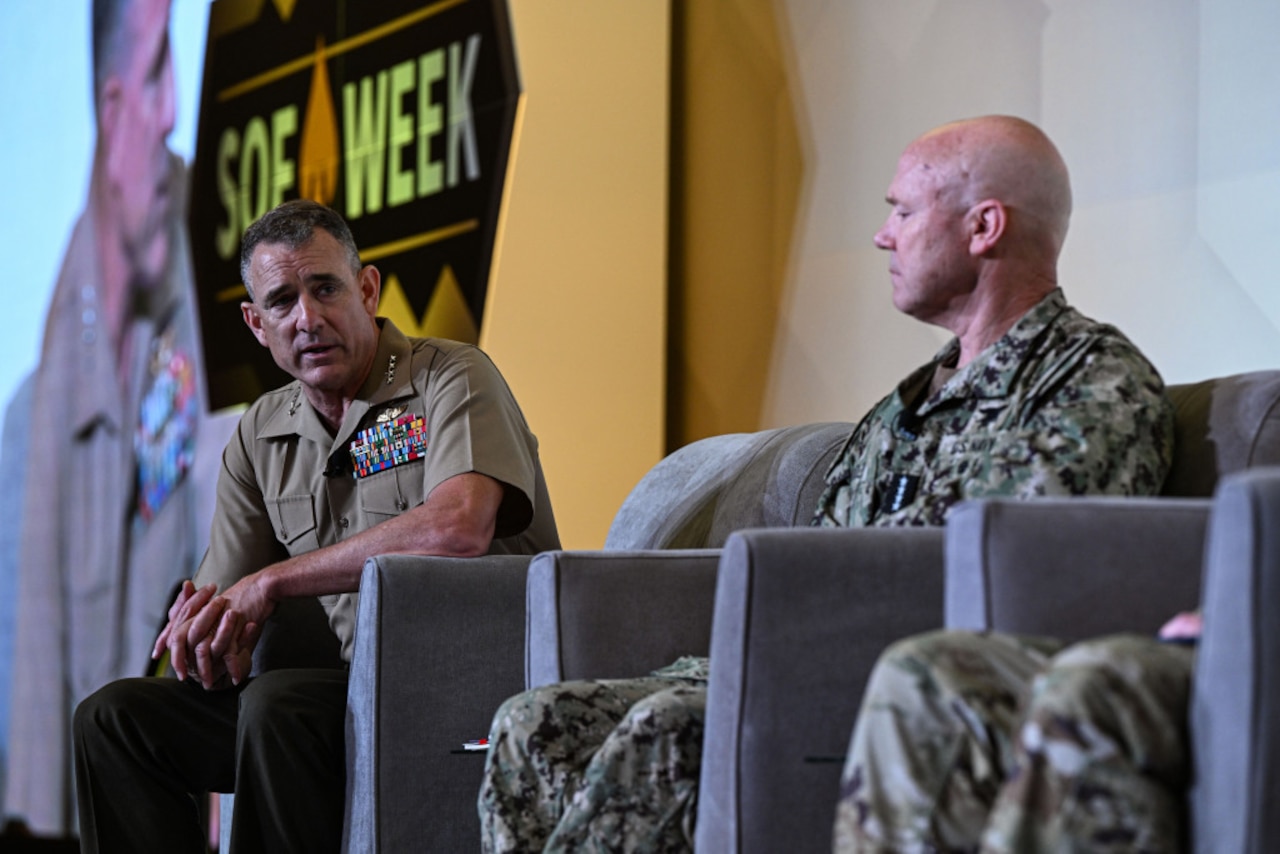 Two men, both wearing camouflage military uniforms, sit next to each other indoors, having a discussion.