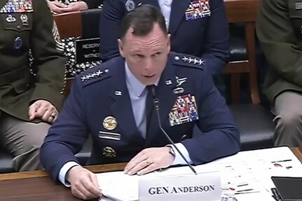 A man in a formal military uniform sits at a desk and speaks into a microphone. In front of him is a place card that reads, "GEN Anderson," and behind him are seated people in similar attire.