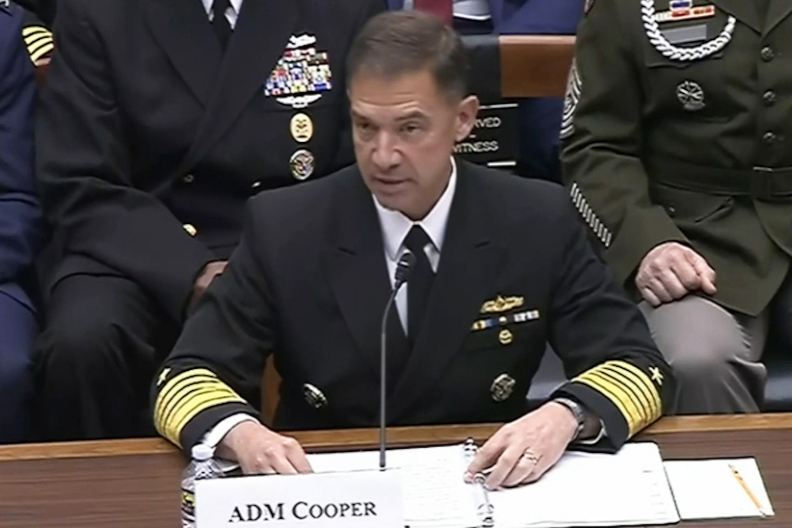 A man in a formal military uniform sits at a desk and speaks into a microphone. In front of him is a place card that reads, "ADM Cooper," and behind him are seated people in similar attire.