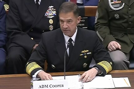 A man in a formal military uniform sits at a desk and speaks into a microphone. In front of him is a place card that reads, "ADM Cooper," and behind him are seated people in similar attire.