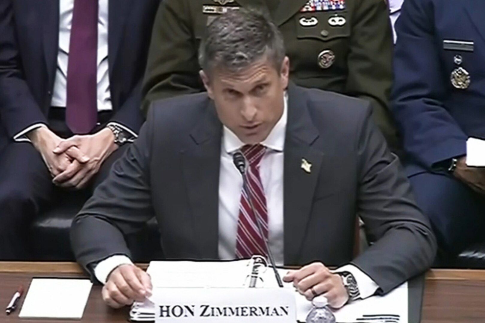 A man in business attire sits at a desk and speaks into a microphone. In front of him is a place card that reads, "HON Zimmerman," and behind him are seated people in similar attire.