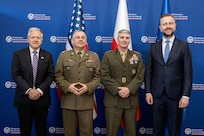 Four men stand side-by-side posing for a photo in front of a blue backdrop with repeating circular logos for the Ministry of National Defence of Poland. From left to right: Mark Brzezinski, the U.S. Ambassador to Poland, wearing a dark suit and striped tie; General Wiesław Kukuła, Chief of the General Staff of the Polish Armed Forces, in an olive-drab Polish army uniform; General Christopher J. Mahoney, Vice Chairman of the Joint Chiefs of Staff, in an olive-drab U.S. Marine Corps uniform; and Władysław Kosiniak-Kamysz, the Polish Minister of National Defence, wearing a dark suit with a blue tie. The United States flag, the Polish flag, and the NATO flag stand behind them.
