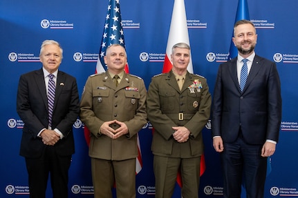 Four men stand side-by-side posing for a photo in front of a blue backdrop with repeating circular logos for the Ministry of National Defence of Poland. From left to right: Mark Brzezinski, the U.S. Ambassador to Poland, wearing a dark suit and striped tie; General Wiesław Kukuła, Chief of the General Staff of the Polish Armed Forces, in an olive-drab Polish army uniform; General Christopher J. Mahoney, Vice Chairman of the Joint Chiefs of Staff, in an olive-drab U.S. Marine Corps uniform; and Władysław Kosiniak-Kamysz, the Polish Minister of National Defence, wearing a dark suit with a blue tie. The United States flag, the Polish flag, and the NATO flag stand behind them.