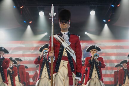 Soldiers dressed in red, Revolutionary War-era uniforms are performing on bugles and other instruments during an indoor performance. In the middle, the soldier is wearing a large furry hat and carrying a long staff with a silver ornamental end. There are several spotlights pointing in different directions from the ceiling, and red and white stripes and other images projecting on a screen on the back wall.