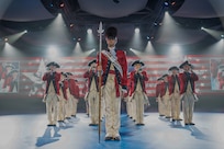 Soldiers dressed in red, Revolutionary War-era uniforms are performing on fifes, bugles and drums during an indoor performance. There are several spotlights in the background, and red and white stripes and other images projecting on a screen on the back wall.