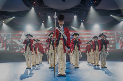 Soldiers dressed in red, Revolutionary War-era uniforms are performing on fifes, bugles and drums during an indoor performance. There are several spotlights in the background, and red and white stripes and other images projecting on a screen on the back wall.