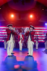 Two soldiers dressed in red Revolutionary War-era uniforms are performing on bugles during an indoor event with colorful lighting. Others are dressed similarly and are performing on other instruments in the background. In the middle, the soldier is wearing a tall furry hat and carrying a long staff diagonally in front of his body.