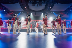 Army soldiers in red Revolutionary War-era uniforms are marching in formation while performing on various musical instruments during an indoor performance with bright spotlights shining down.