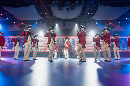 Army soldiers in red Revolutionary War-era uniforms are marching in formation while performing on various musical instruments during an indoor performance with bright spotlights shining down.