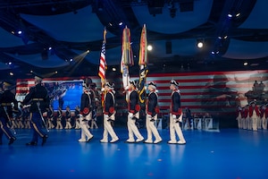 Army soldiers dressed in historic Revolutionary War-era uniforms are marching towards the left while presenting the colors during an indoor show. Dozens of other soldiers in various uniforms are in the background in formations.