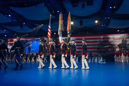 Army soldiers dressed in historic Revolutionary War-era uniforms are marching towards the left while presenting the colors during an indoor show. Dozens of other soldiers in various uniforms are in the background in formations.