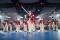 Army soldiers dressed in red Revolutionary War-era uniforms are standing in a formation while performing on various musical instruments. The leader is standing in the front wearing a large furry hat and carrying a long staff diagonally in front of his body.