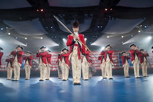 Army soldiers dressed in red Revolutionary War-era uniforms are standing in a formation while performing on various musical instruments. The leader is standing in the front wearing a large furry hat and carrying a long staff diagonally in front of his body.