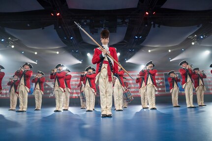 Army soldiers dressed in red Revolutionary War-era uniforms are standing in a formation while performing on various musical instruments. The leader is standing in the front wearing a large furry hat and carrying a long staff diagonally in front of his body.