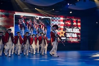Army Soldiers dressed in red, Revolutionary War-era uniforms are marching in formation during an indoor show. The words "U.S. Army Twilight Tattoo" and other graphics appear on the screens in the background.